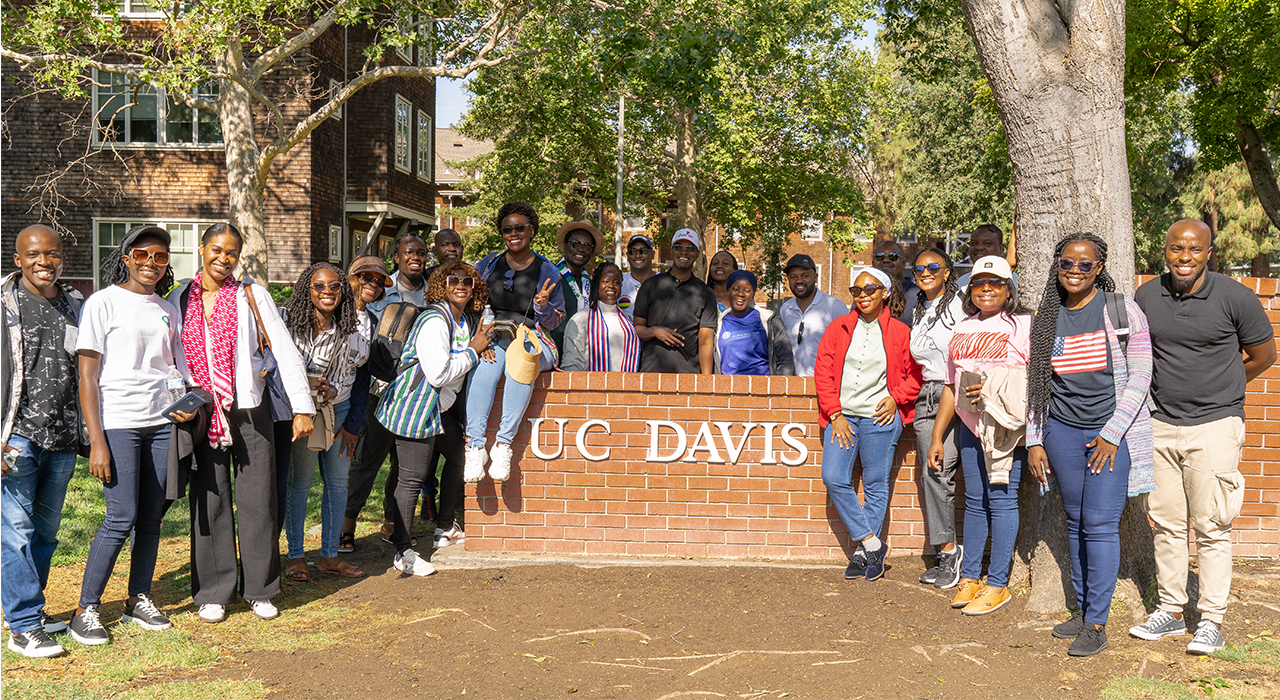 The cohort of 24 Mandela Washington Fellows gathers around a UC Davis sign on a short brick wall on the UC Davis campus.